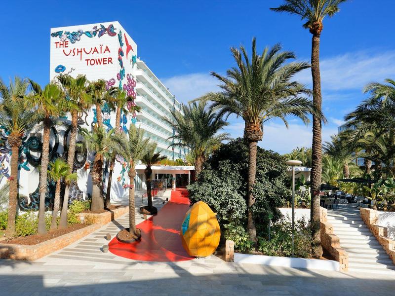 Wejście do hotelu z palmami, białymi schodami i czerwonym chodnikiem pod jasnoniebieskim niebem.