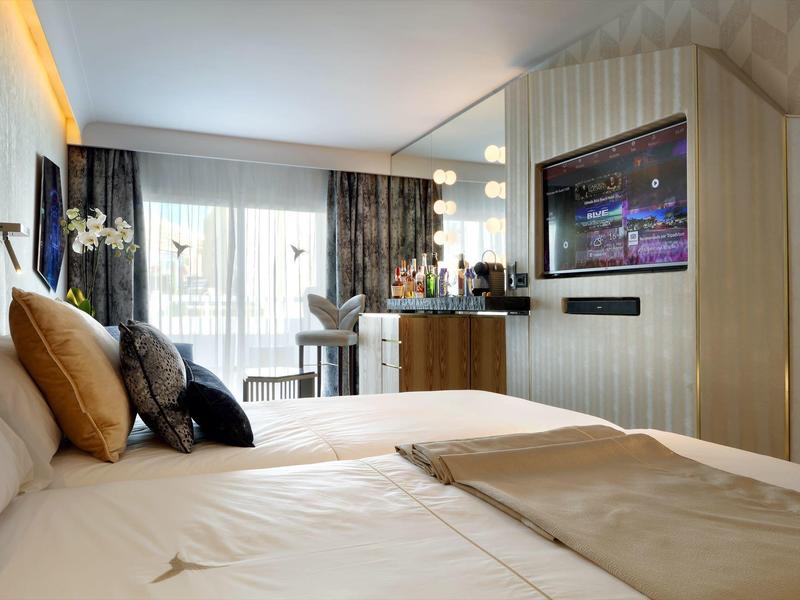 Jasny pokój hotelowy z dużym łóżkiem, telewizorem na ścianie i oknem na balkon.