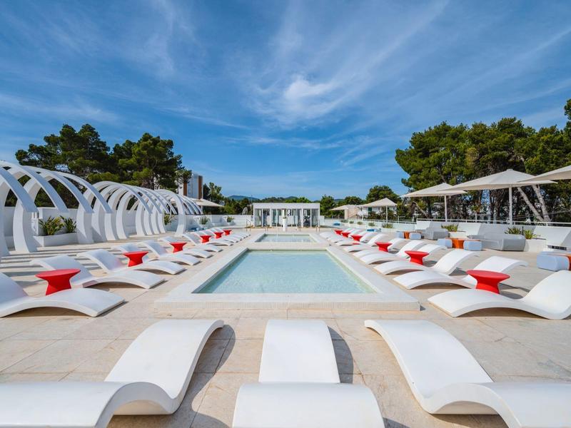 Nowoczesny basen z białymi leżakami i czerwonymi poduszkami pod niebieskim niebem.