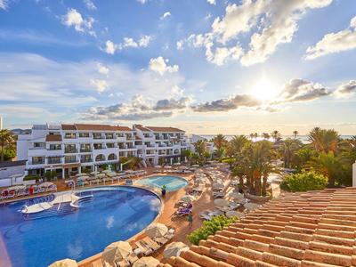 Hotel mit großem Pool, Sonnenterrasse, Palmen und hellem Himmel bei Sonnenuntergang am Meer.