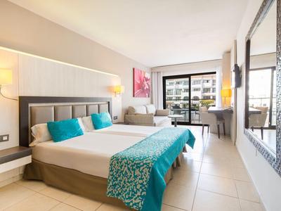 Helles Hotelzimmer mit großem Bett, türkisfarbenen Kissen, Sofa, Schreibtisch und Balkon mit Blick.
