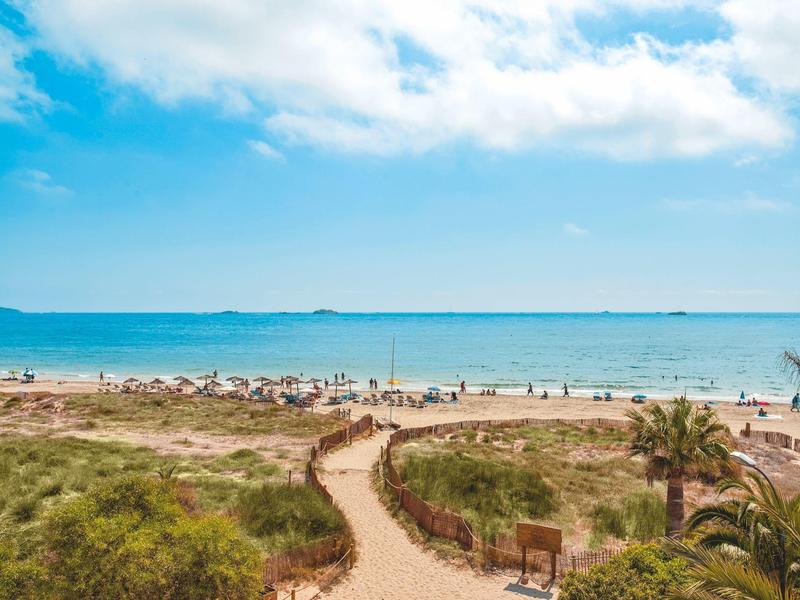Zandpad leidt naar een druk strand met blauwe lucht en kalme zee.