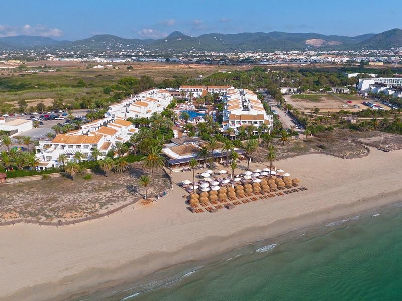 Hotelcomplex met strand, parasols en palmbomen, met bergen op de achtergrond.