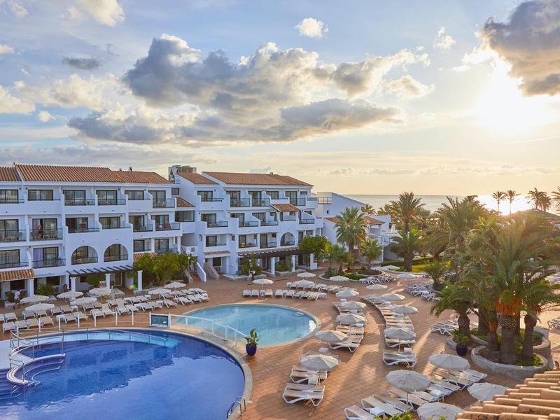 Hotel met zwembaden en ligstoelen onder een bewolkte lucht in de late namiddag.