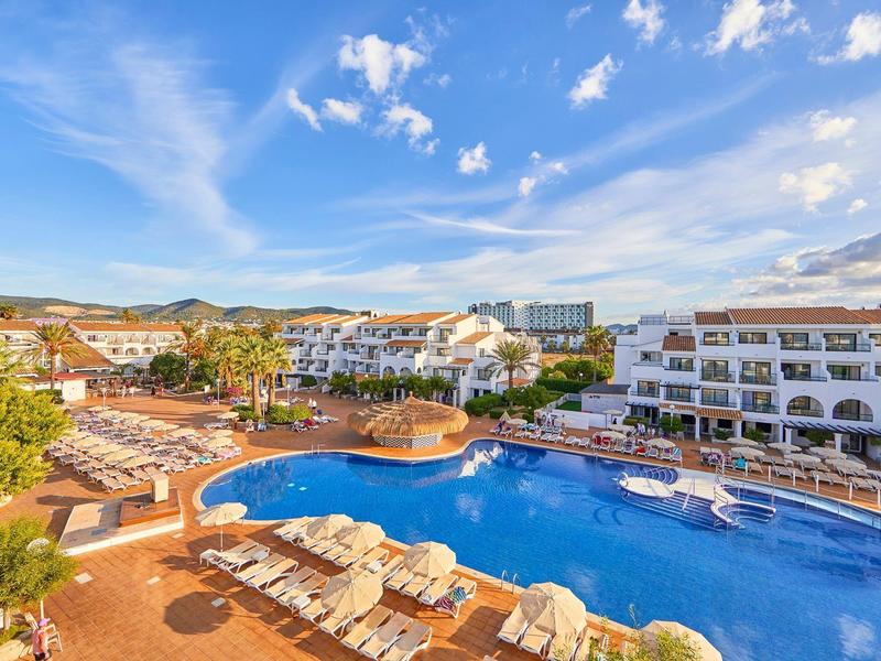 Groot hotelzwembad met ligstoelen en witte gebouwen onder een blauwe lucht.