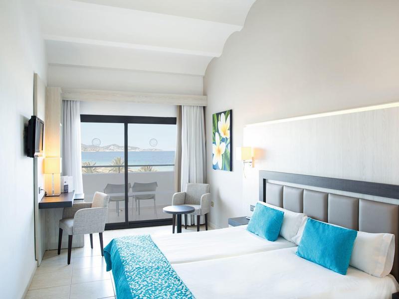 Licht hotelkamer met tweepersoonsbed, blauwe accenten, bureau en balkon met uitzicht op zee.