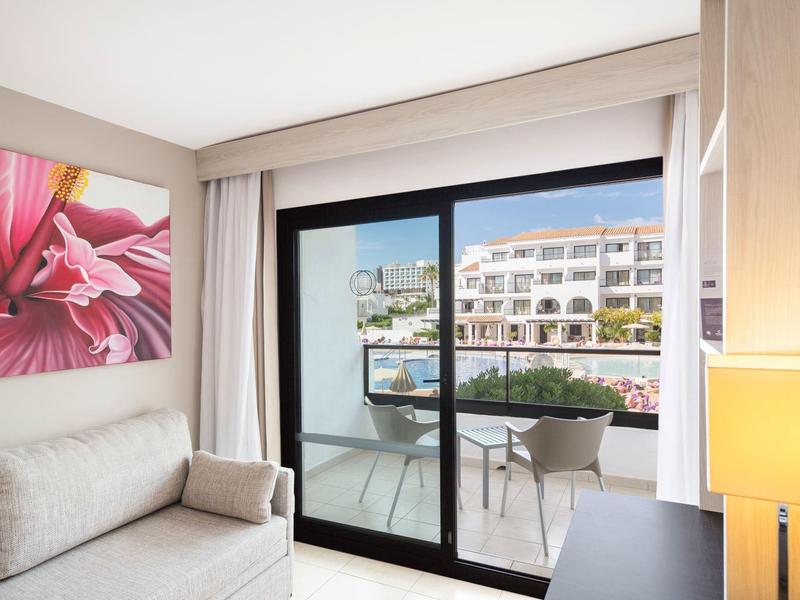 Lichte hotelkamer met bank, grote bloemenschildering en balkon met uitzicht op het gebouw.