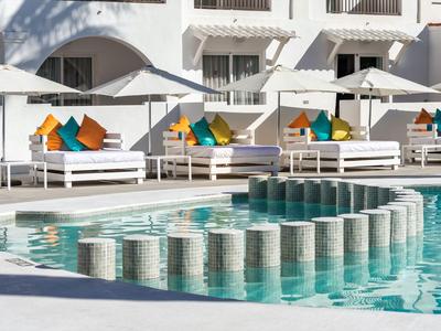 Moderner Poolbereich mit weißen Liegen und bunten Kissen vor einem weißen Hotel.