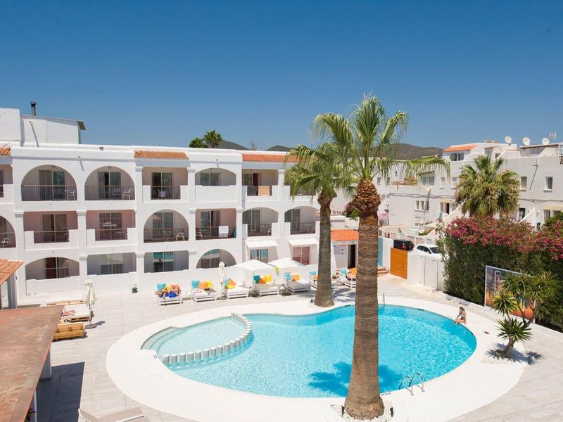 Rond zwembad met zonnig terras voor een wit appartementencomplex onder een blauwe lucht.