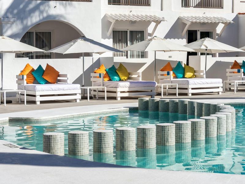 Moderne zwembadzone met witte ligbedden en kleurrijke kussens voor een wit hotelgebouw.
