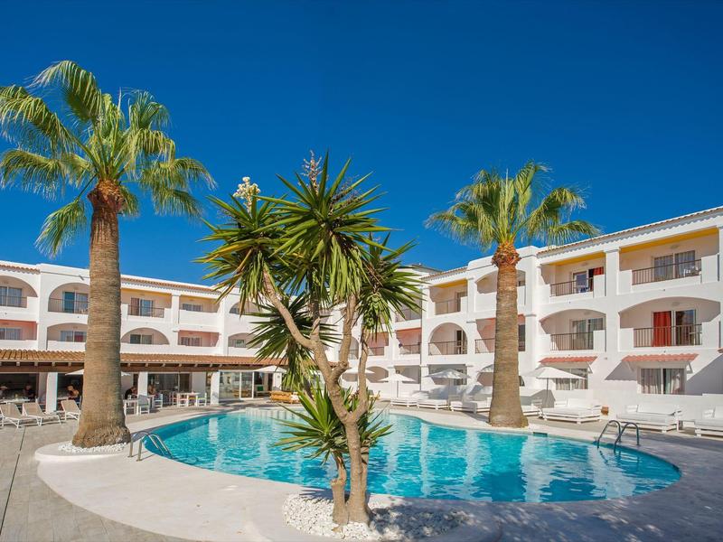 Hotel met ronde buitenzwembad, palmbomen en witte balkons onder een heldere blauwe lucht.