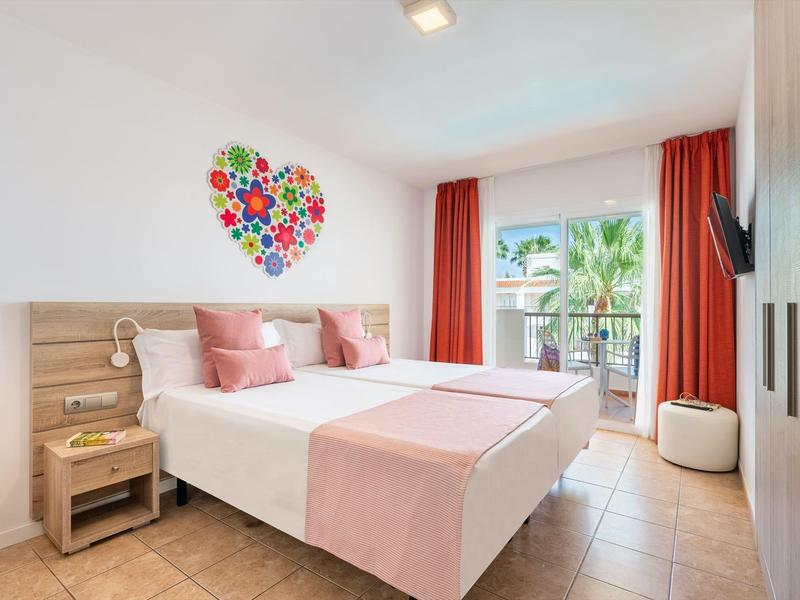 Lichte tweepersoonskamer met roze accenten, hartwanddecoratie en balkon met uitzicht op palmbomen.