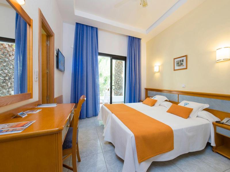 Bright hotel room with double bed, blue-orange curtains, and desk with chair.