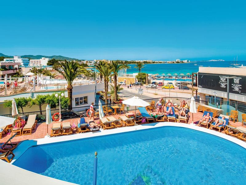 Hotel pool with sun loungers, palm trees, and sea view on a sunny day.