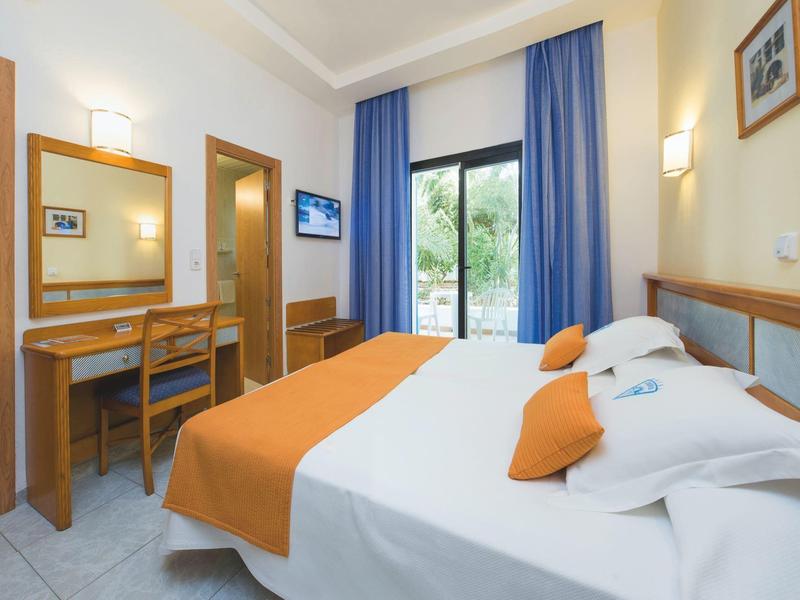Brightly furnished hotel room with double bed, orange accents, and balcony overlooking garden.