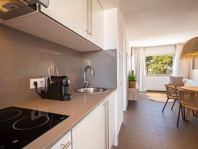 Modern kitchenette with white cabinets, stove, sink, and coffee maker in a bright living space.