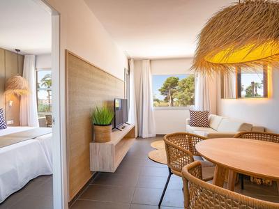 Helles Wohnzimmer mit Rundtisch, Rattanstühlen, moderner Lampe und angrenzendem Schlafzimmer.