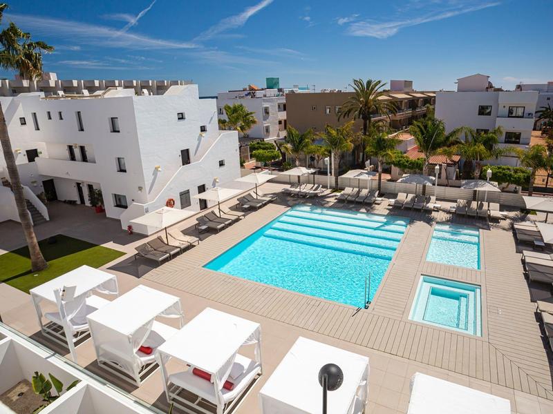 Modern hotel pool with sun loungers and white buildings under a sunny sky.