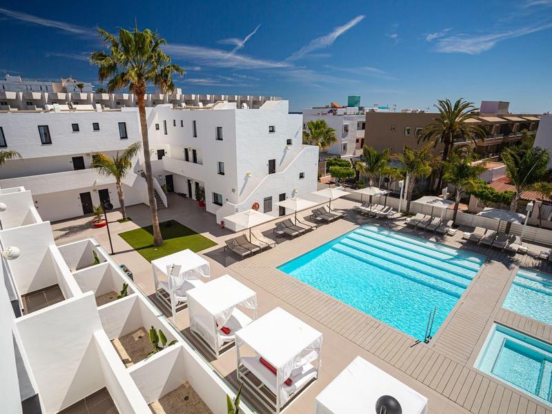 Modern hotel pool with sun loungers and white apartment buildings under a blue sky.