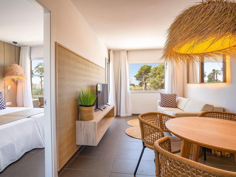 Bright hotel room with dining area, wicker chairs, and adjoining bedroom with large bed.