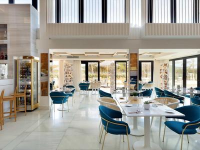 Modern hotel dining area with blue chairs, white tables, and large windows letting in natural light.