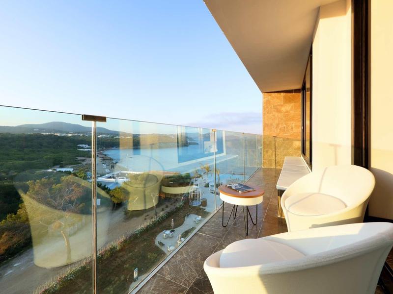 Balcony with white chairs overlooking a coastal road and hills under a clear sky.