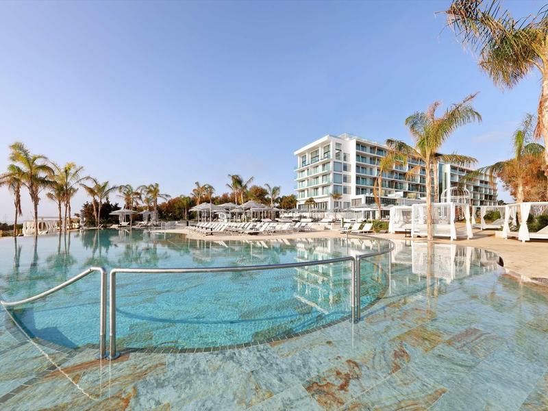 Modern hotel with a large outdoor pool and palm trees under a clear blue sky.