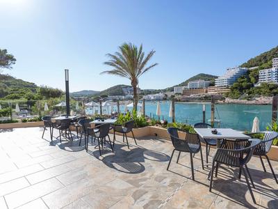 Terrasse mit Tischen und Stühlen mit Blick auf eine Bucht und Hotelgebäude.