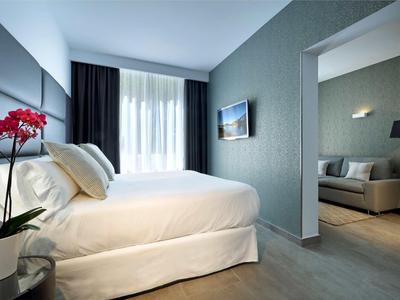 Moderní hotelový pokoj s bílou postelí, tmavými závěsy a viditelným obývacím koutem.