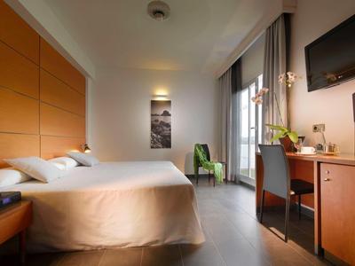 Moderní hotelový pokoj s manželskou postelí, světlým dřevěným nábytkem a velkým oknem s závěsy.