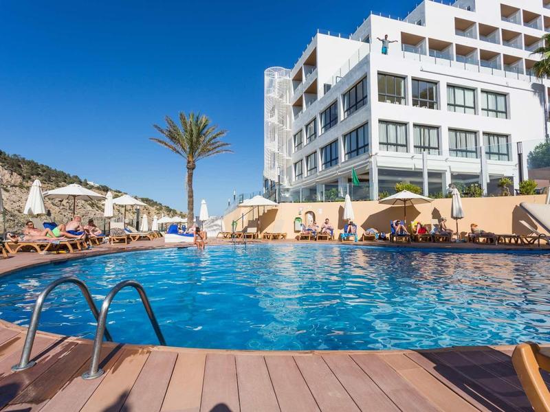 Modrý bazén s lehátky a moderním hotelem pod jasnou oblohou.