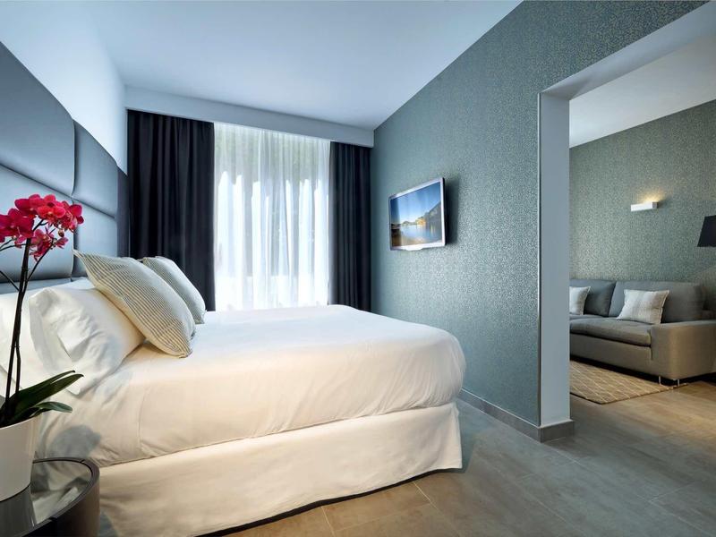 Moderní hotelový pokoj s bílou postelí, tmavými závěsy a viditelným obývacím koutem.