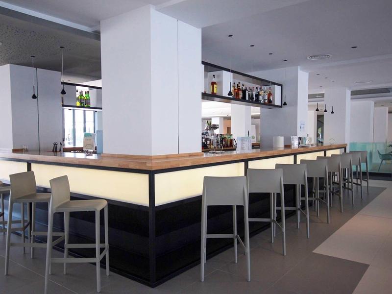 Moderne hotelbar met verlichte toonbank en grijze barkrukken in een lichte omgeving.