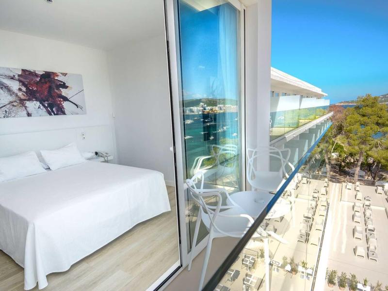 Licht hotelkamer met balkon met uitzicht op zwembad, ligstoelen en de zee.