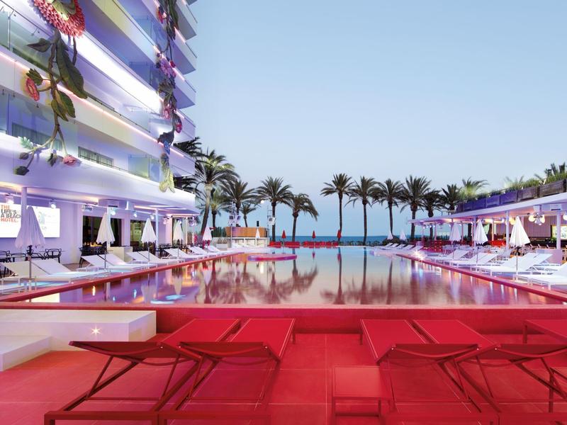 Moderní hotelový bazén s červenými lehátky a palmami pod modrou oblohou.