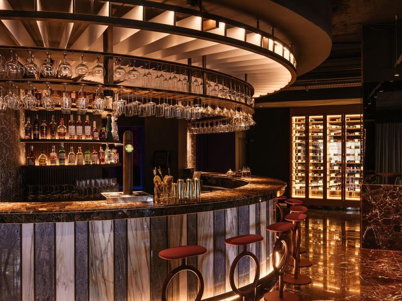 Elegantní hotelový bar s kulatým pultem, stylovými barovými židlemi a teplým osvětlením.