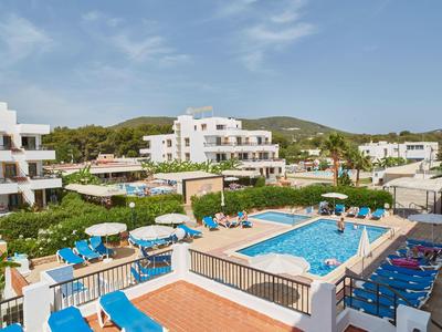 Hôtel avec piscine extérieure, chaises longues et vue sur des collines vertes sous un ciel bleu.