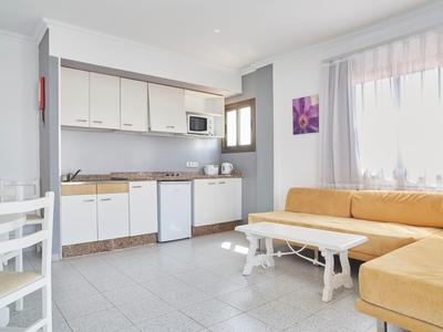 Salon lumineux avec canapé d'angle jaune, kitchenette blanche et table à manger avec chaises.