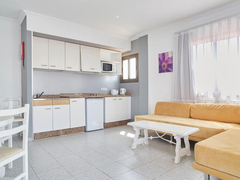 Salon lumineux avec canapé d'angle jaune, kitchenette blanche et table à manger avec chaises.