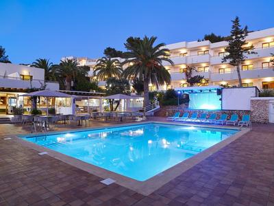 Osvětlený venkovní bazén před vícepodlažním hotelem za soumraku s palmami a posezením.