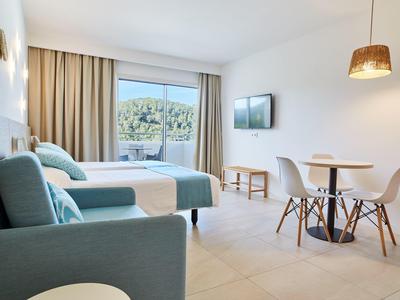 Světý hotelový pokoj se dvěma postelemi, pohovkou, jídelním stolem a balkonem s výhledem do zeleně.