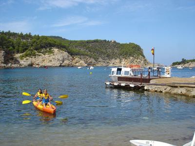 Dva lidé pádlí v oranžovém kajaku na klidné vodě u mola a zalesněného pobřeží.