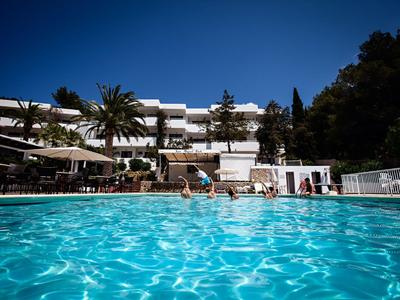 Čirý modrý bazén před bílou hotelovou budovou s palmami a slunečnou oblohou.