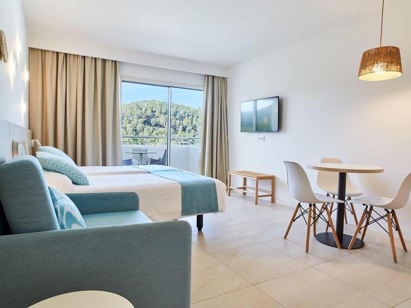 Světý hotelový pokoj se dvěma postelemi, pohovkou, jídelním stolem a balkonem s výhledem do zeleně.