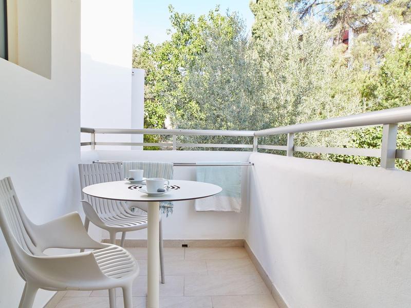 Malý balkon s bílým kulatým stolem, dvěma židlemi a výhledem na zelené stromy.