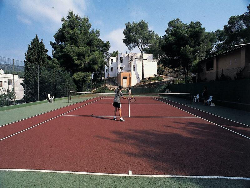 Tenisový kurt s červeným povrchem obklopený stromy za jasného denního světla.