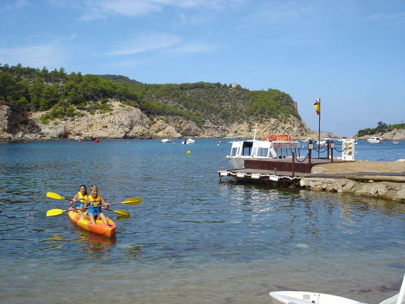 Kajakáři veslující na klidné vodě poblíž skalnatého pobřeží a zalesněných kopců pod jasnou oblohou