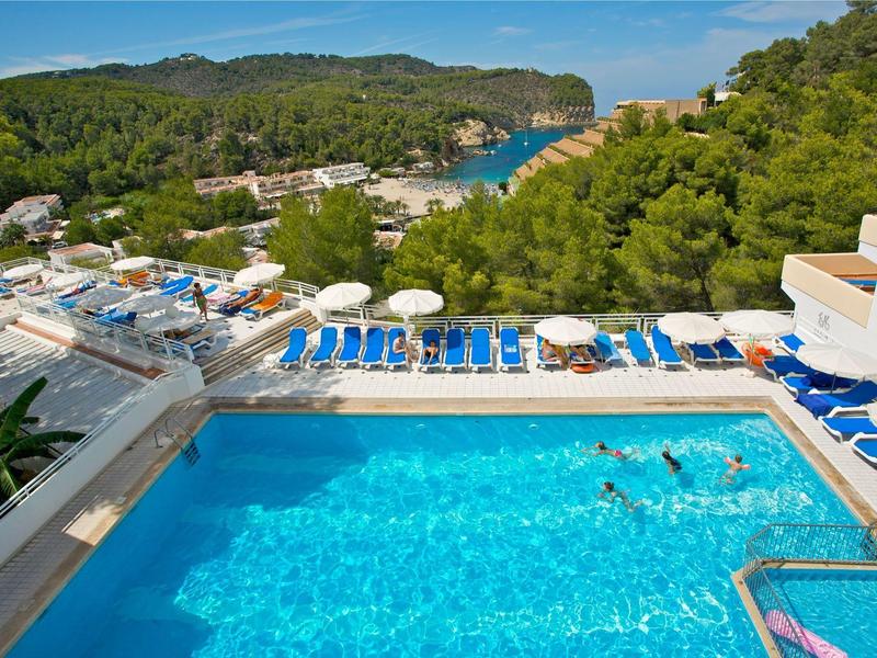 Pohled na venkovní bazén s modrými lehátky a zalesněnými kopci v pozadí.