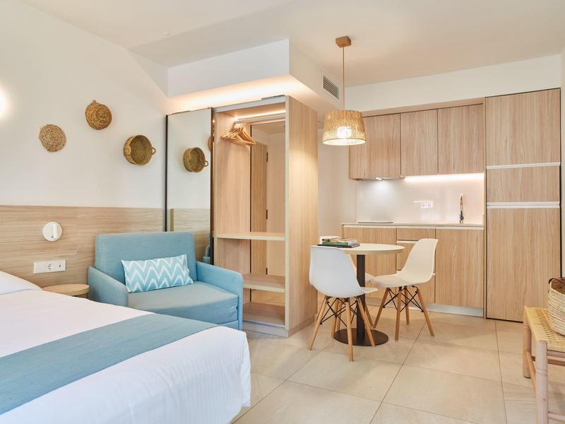 Moderní hotelový pokoj s postelí, pohovkou, stolem a kuchyňským koutem v světlých barvách.