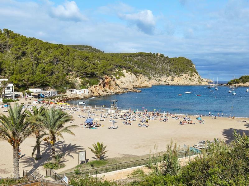 Kamenitá pláž s návštěvníky, obklopená zelenými kopci a několika budovami u moře.